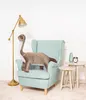 A plush dinosaur toy is placed on a light blue armchair in a room with a floor lamp and side tables. Náhled obrázku produktu