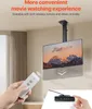 A hand holding a remote control is shown in the foreground, with a TV mounted on a motorized ceiling bracket in the midground. The TV displays a mountain landscape. A wired controller is visible in the bottom right corner. Podgląd zdjęcia produktu