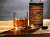 A glass of whiskey with ice sits next to a bottle of Shanky's Whip Original Black Irish Whiskey Liqueur on a wooden surface. Produktbild-Vorschau