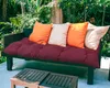 A bench cushion and pillows are arranged on a dark bench in an outdoor setting with lush greenery in the background. Produktbild-Vorschau