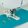 A close-up view of a sewing machine's buttonhole foot attachment, demonstrating its use on teal fabric with yellow markings. Produktbild-Vorschau