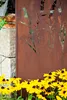 A decorative metal screen with a floral cut-out design stands next to a stone wall, with yellow flowers in the foreground. Produktbild-Vorschau