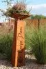 A decorative metal pillar with a planter on top, set in a garden with tall grasses and gravel. Produktbild-Vorschau