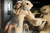 A woman is sitting on a chair next to a large teddy bear at a table. Another large teddy bear is visible in the background. Náhled obrázku produktu