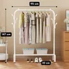 A white metal garment rack with clothes hanging on it, placed in a room with a light beige wall and wooden floor. There are two storage bins on the bottom shelf and shoes on the floor. Náhled obrázku produktu