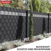 A black PVC privacy screen fence installed in a garden setting with a tall cypress tree and some low-lying plants in the foreground. Produktbild-Vorschau