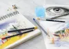 Art supplies laid out on a table, including sketchbooks, colored pencils, a brush, and a glass of water. Produktbild-Vorschau