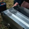A person is holding a stainless steel grill attachment over a grassy area, with a gas canister in the background. Produktbild-Vorschau