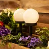 Two white globe solar lamps are placed in a planter box with purple violets and green foliage. Produktbild-Vorschau