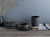 A set of black stoneware dishes and bowls are arranged on a wooden table, with a vase of dried branches in the background. Náhľad obrázku produktu