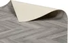 A close-up, angled view of a roll of vinyl flooring with a herringbone wood pattern. The edge of the roll is lifted, revealing the beige backing. Produktbild-Vorschau