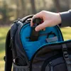 A hand is placing a small black digital camera into the open compartment of a gray backpack. The backpack has a bright blue interior. Produktbild-Vorschau