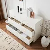 A white sideboard with open drawers filled with folded clothes and other items, placed on a wooden floor next to a rug and a decorative plant in a vase. Anteprima dell’immagine prodotto