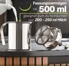 A stainless steel milk frother with its lid next to it, with milk being poured into it. The background is dark and shows a counter with wooden elements. Produktbild-Vorschau