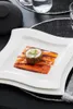 A white square plate with wavy edges, holding a single sushi roll on top of thinly sliced orange vegetables. The plate is on a black surface with a black placemat and a black teapot in the background. Produktbild-Vorschau