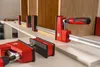 Several red and silver clamps are holding together wooden panels on a white surface. Produktbild-Vorschau