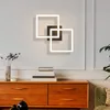 A modern wall lamp with geometric square shapes mounted on a light gray wall above a wooden dresser. Produktbild-Vorschau