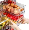 A person's hands are shown pulling out a clear plastic organizer bin filled with vegetables from a refrigerator. Náhled obrázku produktu