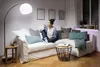 A woman sits on a couch in a living room, holding a smartphone and looking at it. The room is dimly lit, with a floor lamp providing light. There is also accent lighting on a shelf. Produktbild-Vorschau