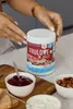 A person's hands are holding a jar of ALLNUTRITION FRULOVE In Jelly Cherry, with bowls of jam, nuts, and cream in the foreground. Produktbild-Vorschau