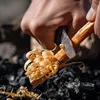 A close-up of a hand using a knife to shave wood shavings from a stick over a pile of charcoal. Produktbild-Vorschau