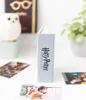 A small, light blue Harry Potter photo album stands upright on a white surface, with several photos scattered around it. In the background, a white owl figurine and a potted plant are visible. Anteprima dell’immagine prodotto