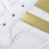 A close-up view of a white Adidas shoe with gold stripes. Produktbild-Vorschau