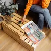 A person's hand is reaching for a cup of coffee on a wooden crate coffee table, with magazines open on the table. Náhľad obrázku produktu