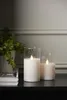 Two LED candles in glass holders sit on a dark surface next to a potted plant. Podgląd zdjęcia produktu