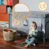 A child is sitting on the floor next to a baby travel cot, playing with a toy xylophone. The cot has a mesh side with arrows indicating airflow, and a close-up inset shows the mesh detail. A basket of toys is also visible. Anteprima dell’immagine prodotto