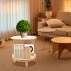 A small, round, wooden side table with books and a plant on it, placed in a living room setting with a sofa and curtains in the background. Náhled obrázku produktu