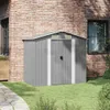 A grey metal garden shed is placed next to a brick house with a window. The shed is surrounded by green grass and a stone pathway. Podgląd zdjęcia produktu