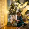A woman is sitting on the floor next to a decorated Christmas tree, placing a gift into a gift bag. Anteprima dell’immagine prodotto