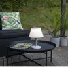 A solar LED table lamp is placed on a black outdoor coffee table next to a magazine, with a cushion on a chair in the background and potted plants to the right. Produktbild-Vorschau