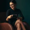 A woman sits in a brown leather armchair, holding a black coffee cup. Produktbild-Vorschau
