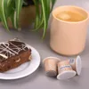 A slice of chocolate cake with white icing, a cup of coffee, and three coffee capsules are arranged on a gray surface next to a potted plant. Produktbild-Vorschau