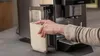 A person's hand is shown holding a clear milk container filled with milk, placing it into a coffee machine. Anteprima dell’immagine prodotto