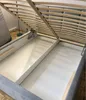 The image shows the inside of a bed frame with the mattress support slats lifted, revealing the storage compartment underneath. The bed frame is upholstered in gray fabric. Náhled obrázku produktu