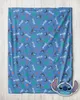 A blue fleece blanket with a repeating pattern of Stitch characters and the word 'Stitch' is laid out on a light wooden surface. A cutout of Stitch's head is placed in the bottom right corner. Produktbild-Vorschau