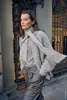 A woman walks past a building with ornate metal gates, wearing a grey knitted cardigan and trousers. Produktbild-Vorschau