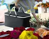 A person is at a flower market, holding a black picnic basket and reaching for flowers. Produktbild-Vorschau