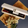 A white wooden wine box with a bottle of red wine inside, placed on a wooden board with a bouquet of white flowers in the foreground. Produktbild-Vorschau