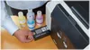 A person is shown inserting a black ink cartridge into a printer while four bottles of ink (yellow, blue, pink, and white) are lined up next to it. Produktbild-Vorschau