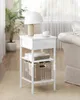 A white nightstand with a lamp, cup, books, and a basket on it, placed in a room with a plant and part of a couch visible. Náhled obrázku produktu