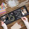 A person's hands are shown decorating a black scrapbook with photos, stickers, stencils, and metallic pens. The scrapbook is open, displaying a couple's photo on the left page and a baby photo on the right page. Various craft supplies are scattered around  Produktbild-Vorschau