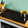 A black induction cooktop with a pot of spaghetti on it, set on a wooden countertop with a knife block and a bowl of tomatoes in the background. Produktbild-Vorschau