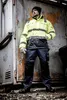 A man wearing workwear stands in front of a weathered industrial container. Produktbild-Vorschau
