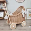 A wicker doll carriage with wooden accents and a large bow decoration, placed on a wooden floor next to a shelf with various items. Anteprima dell’immagine prodotto