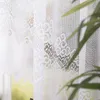 A close-up view of a white lace curtain with a floral pattern, partially obscuring a window with greenery visible outside. Náhled obrázku produktu