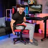 A man is sitting in a red and black gaming chair in a room with gaming equipment. Produktbild-Vorschau